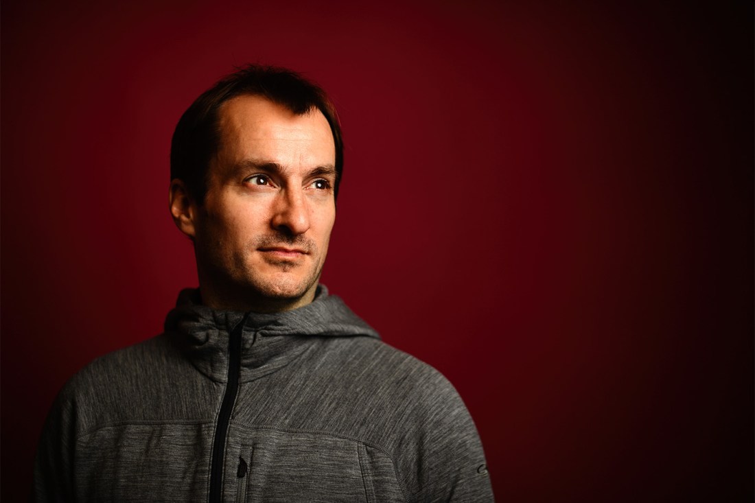 Portrait of Aron Stubbins, wearing an athletic jacket in front of a red background.