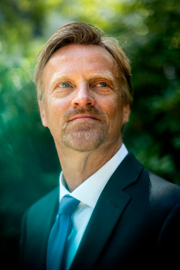 A man in suit and tie looks off to the right, standing in front of a foliage background.