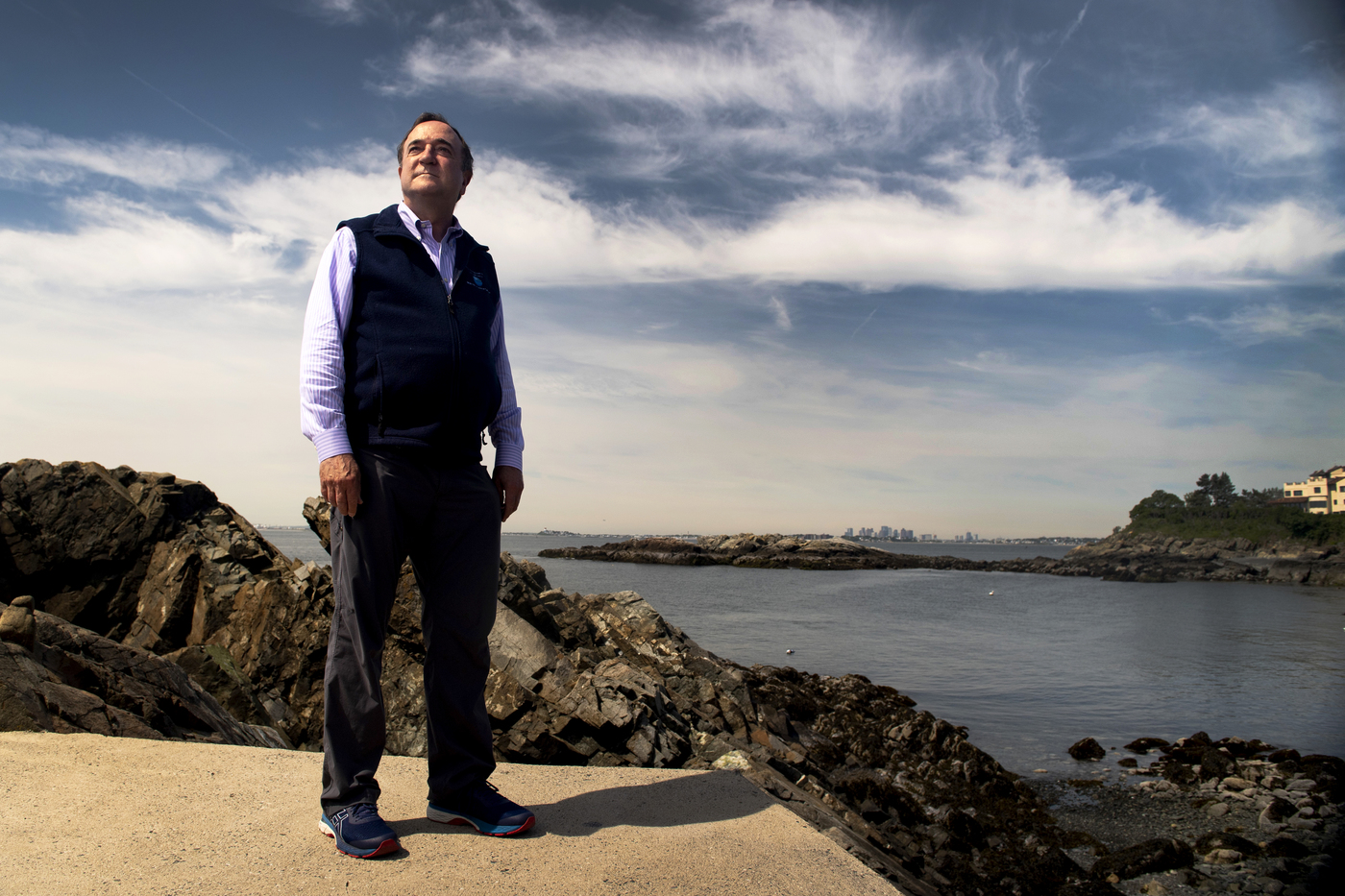 William Detrich Professor of Marine and Environmental Sciences. A man stands by the rocky ocean shore under a blue sky with wispy clouds, with the city skyline in the background.