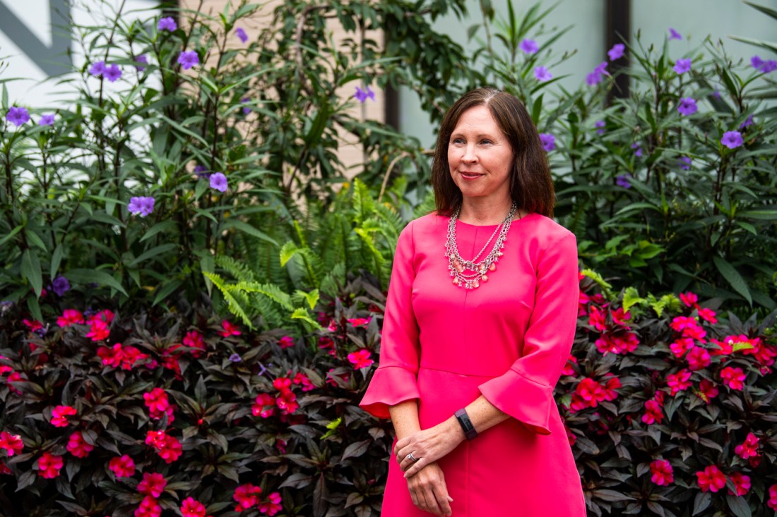 Amy Farrell wearing a pink dress standing in front of bushes with pink and purple flowers on them. 