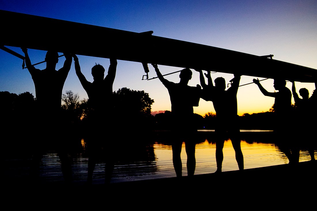 Crew members hold a rowing boat next to a river at sunset.