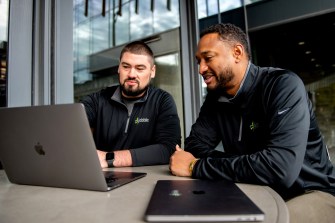 Two people sit next to each other in front of a laptop computer.