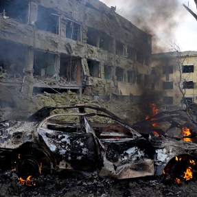 A mangled, burning car in front of war-damaged buildings