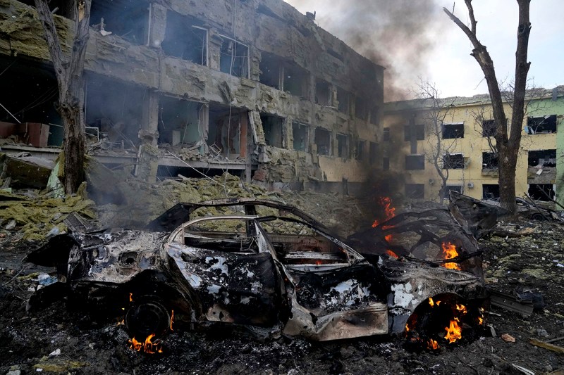 car burning and smoking in front of a destroyed building