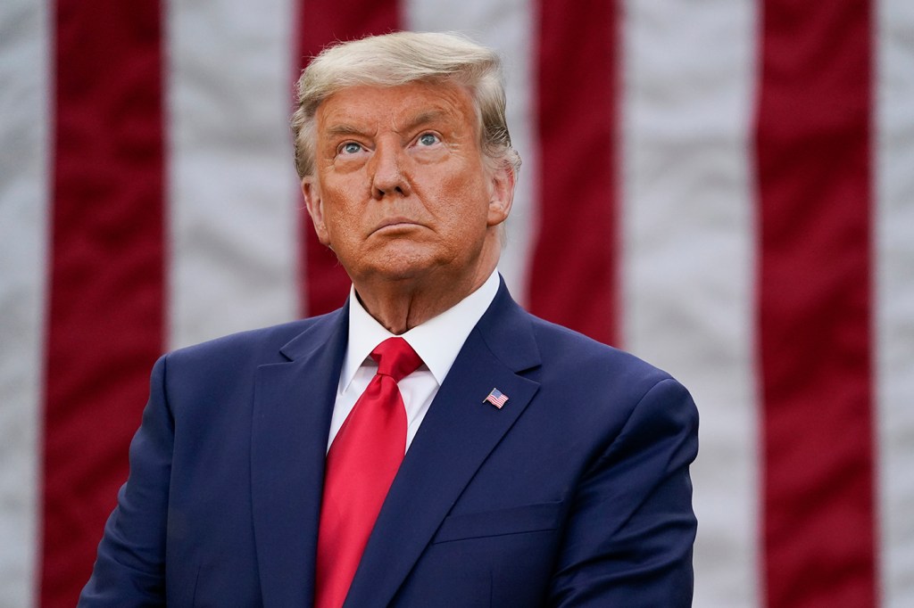 tan old man wearing blue suit and red tie standing in front of american flag