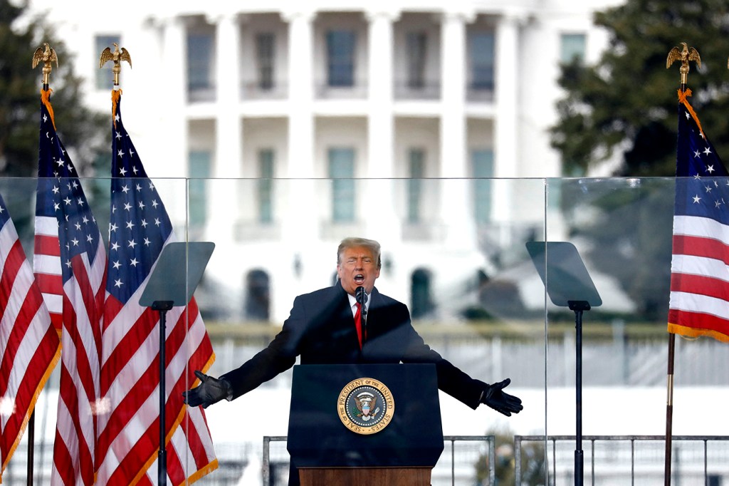 man wearing black coat and gloves stands at pulpit with arms widespread surrounded by us flags