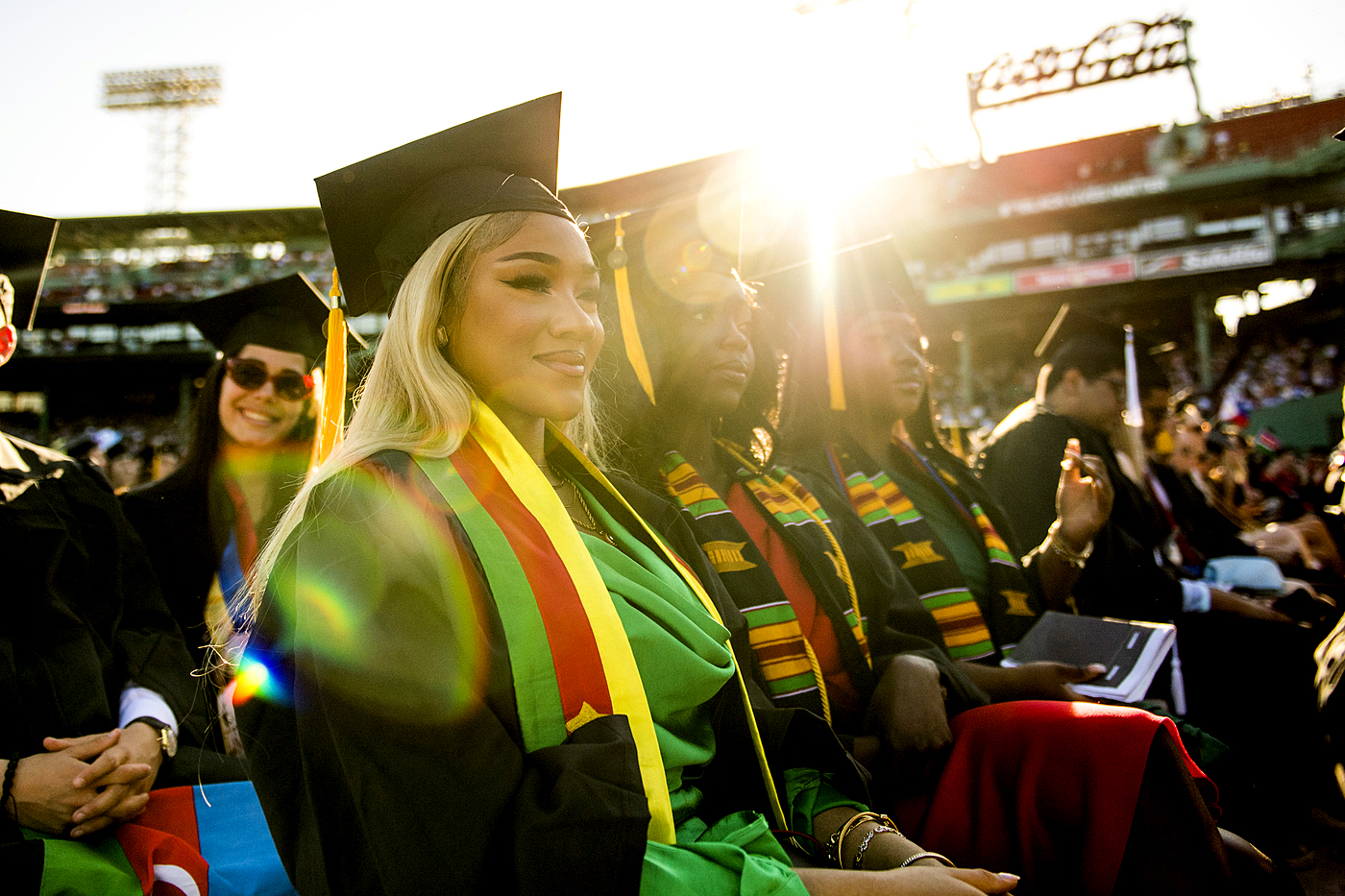 graduates sit in fenway stadium with the sun behind them