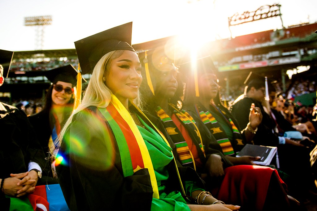 graduates sit in fenway stadium with the sun behind them