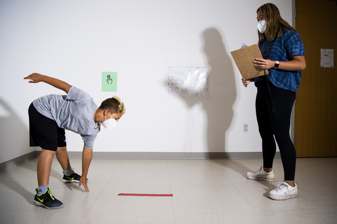 A study participant performs an exercise by bending over and touching the floor with one hand as a researcher holding a clipboard looks on  