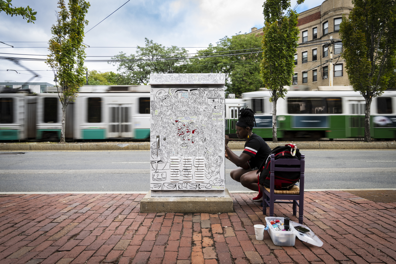 A person wearing a black shirt is doing a mural painting a large utility box outside. 