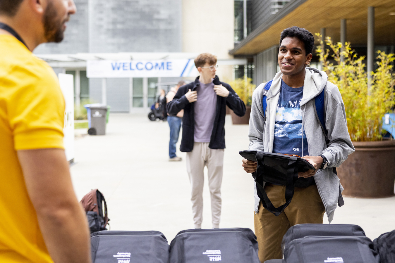 A student smiles as he stands on a sidewalk with a welcome sign and other passersby seen in the background