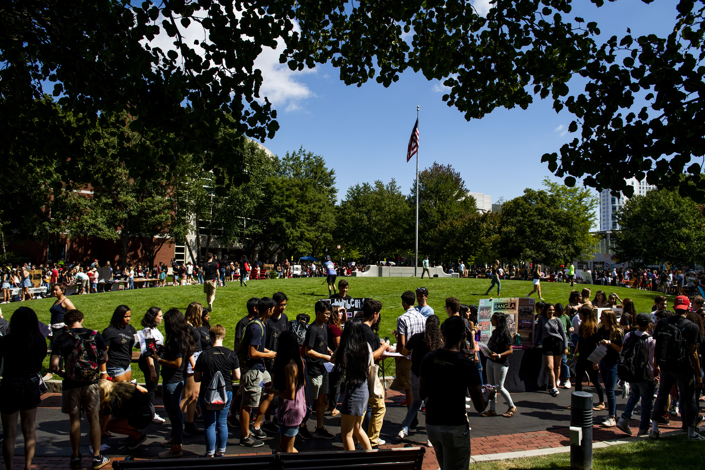 People walk around a large patch of grass outside on a sunny day.