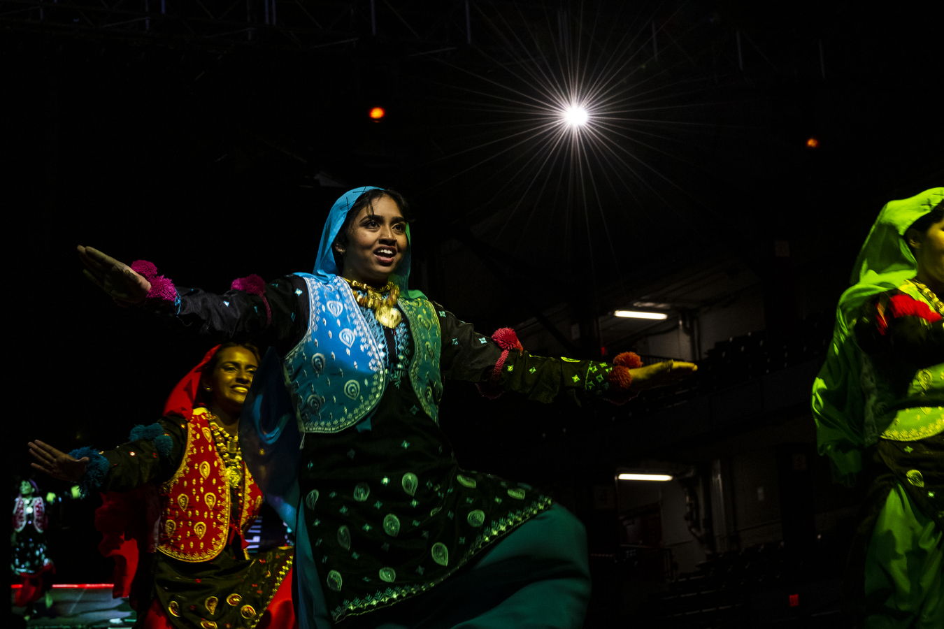 Three people are wearing multi-colored outfits on a dimly lit stage. 