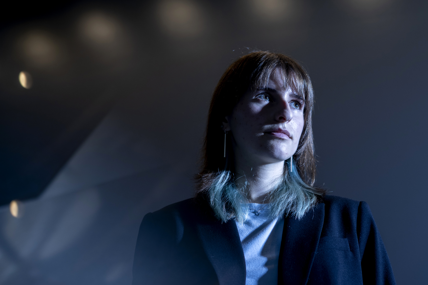 A person wearing a black blazer and grey sweater is standing in a dimly lit room. 