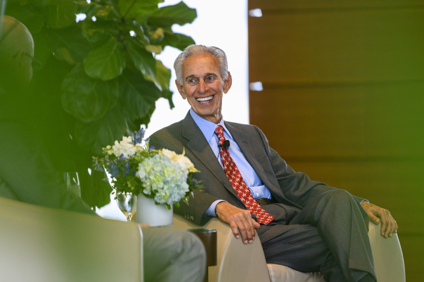 A person wearing a red tie, blue buttoned shirt, and grey suit is laughing. 