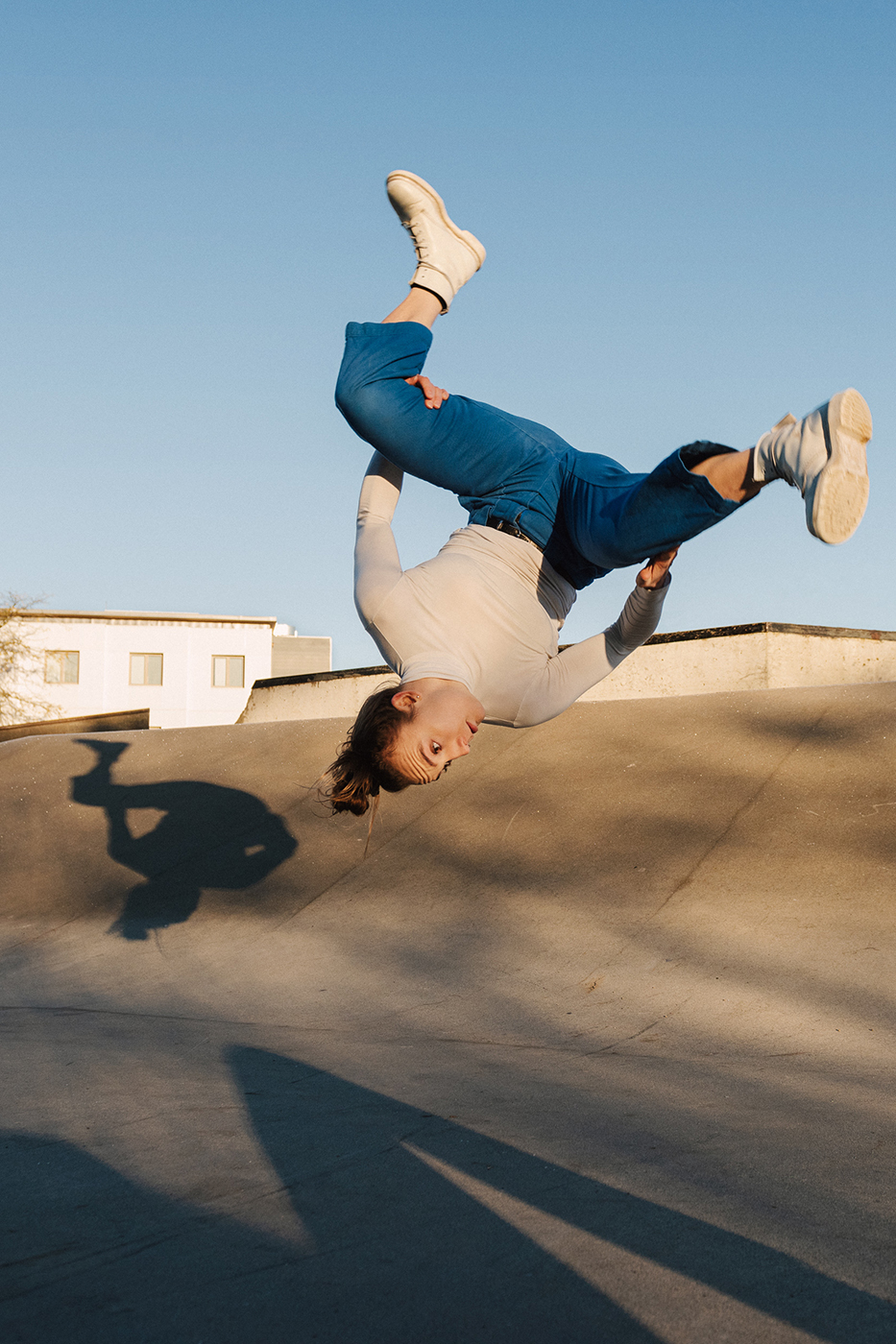 A person wearing a white long-sleeved shirt and jeans is dancing on the pavement in the sunlight. 