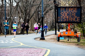 People are running outside near a large sign with orange text over a black background: "Please social distance."