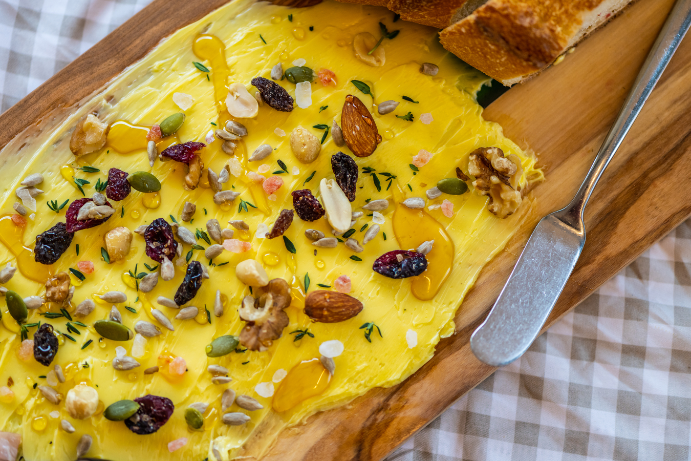 A butter board covered in nuts, honey, and seeds on a wooden board with a butter knife. 