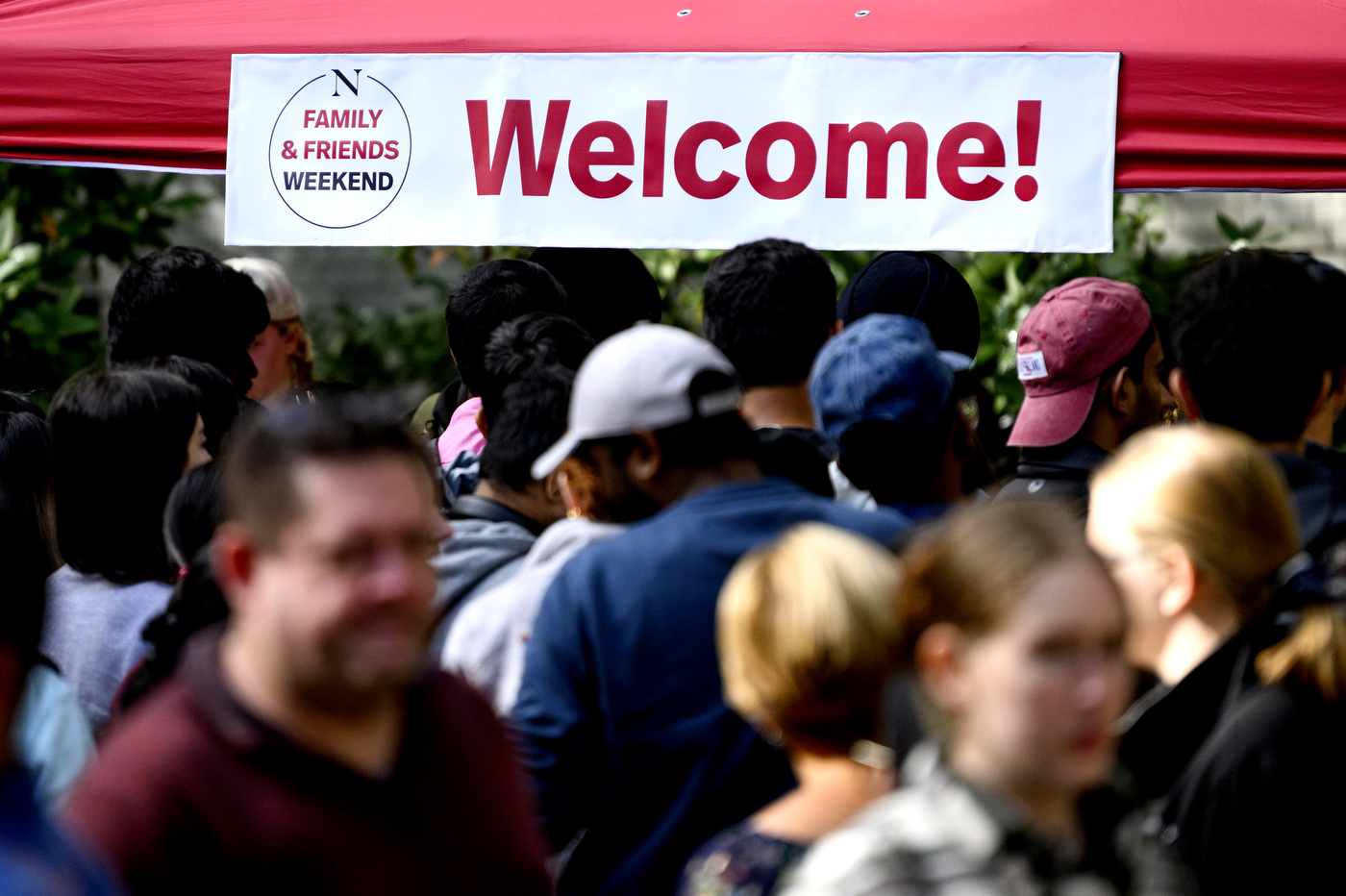 People are walking by a red tent and a white sign with red text: "Welcome! Family & Friends Weekend".