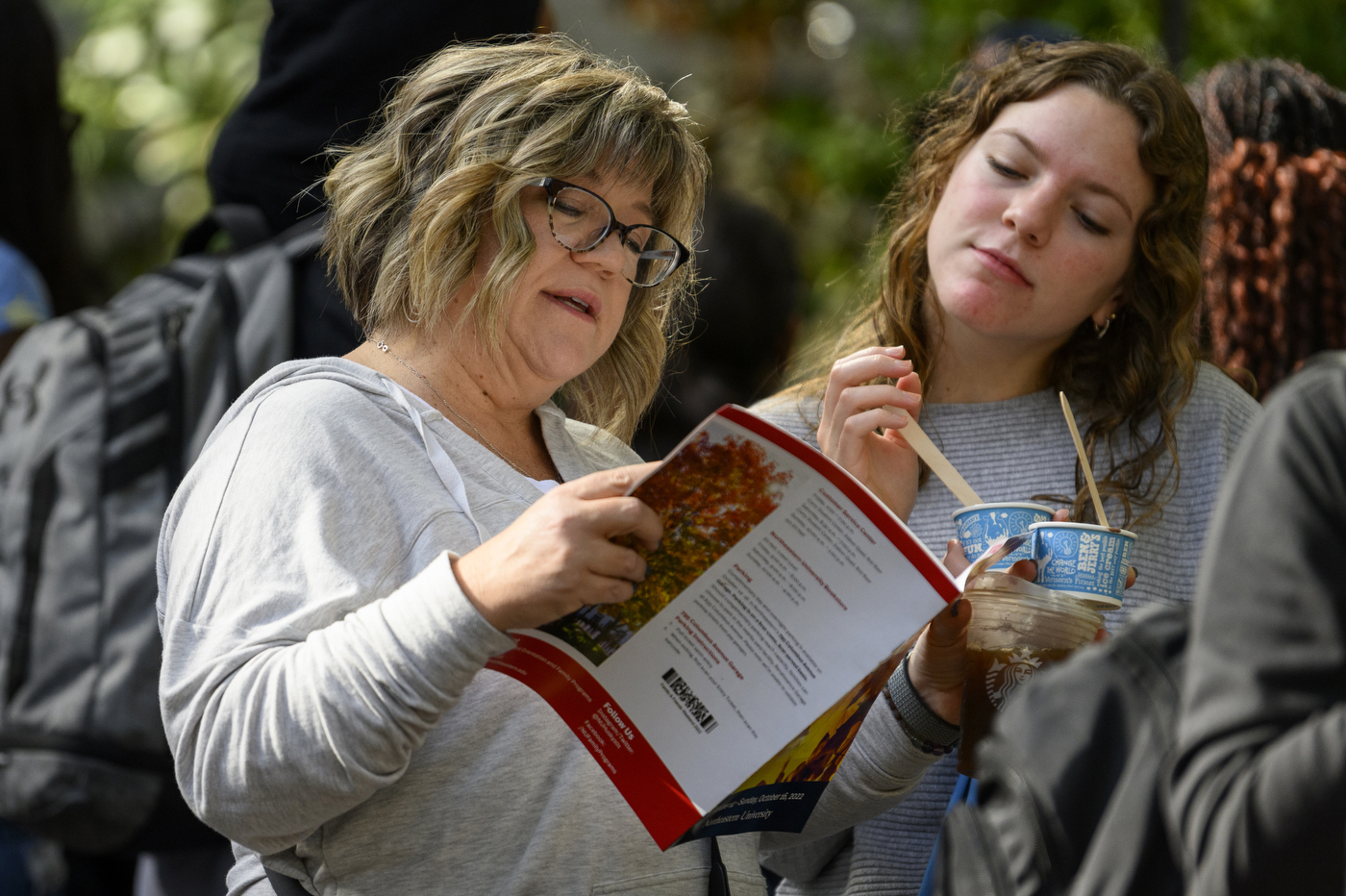 A person holding a book stands next to another person eating ice cream.