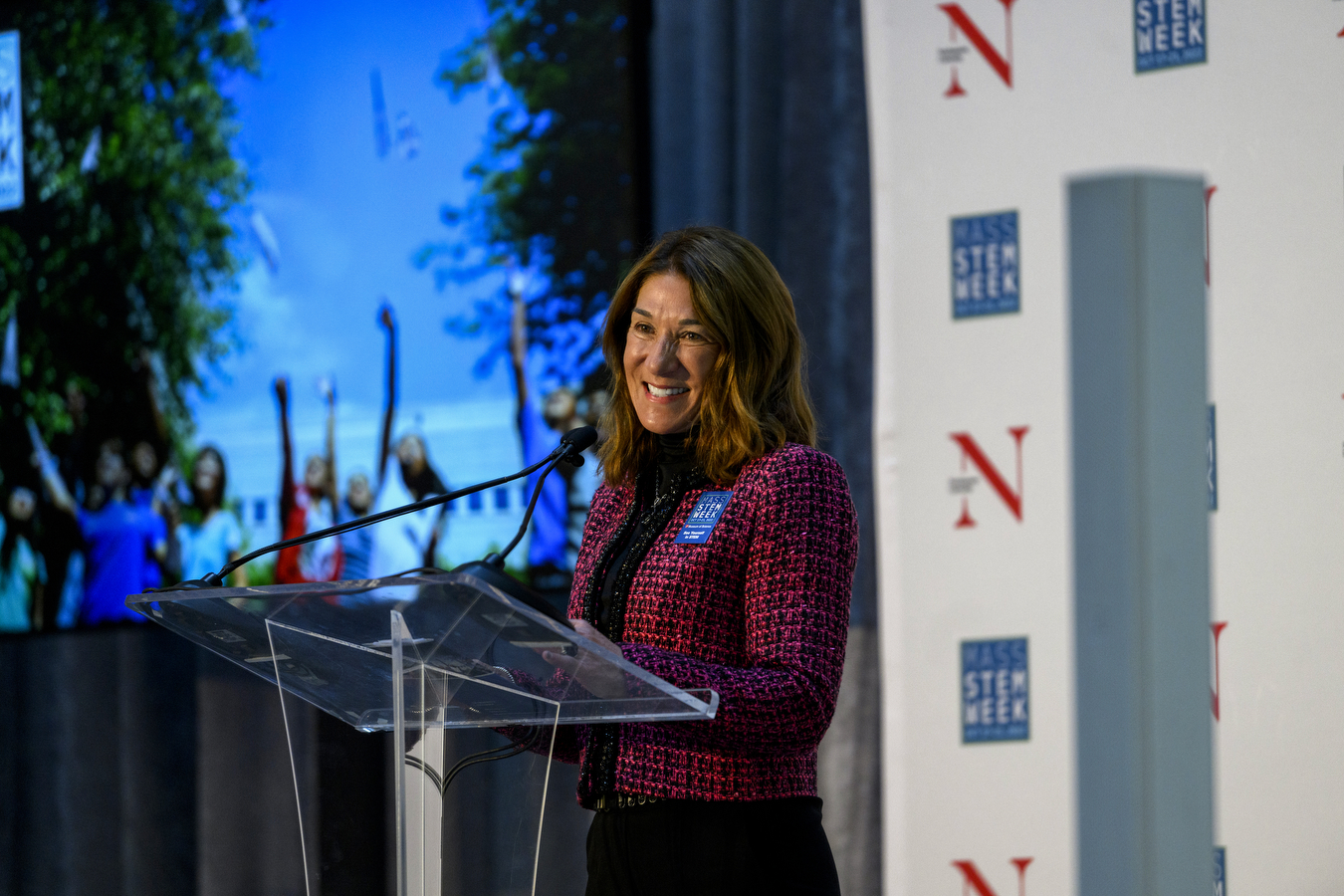 A person wearing a red jacket and black outfit stands at a glass podium on a stage celebrating Stem Week 2022. 