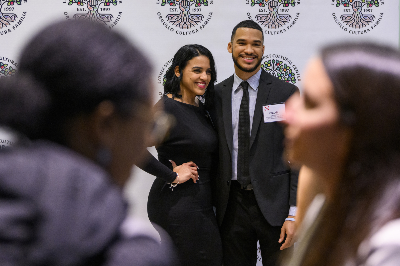 Two participants posing for a photo in front of a display with the Latinx Student Cultural Center logo. The bottom of the logo says orgullo cultura familia.