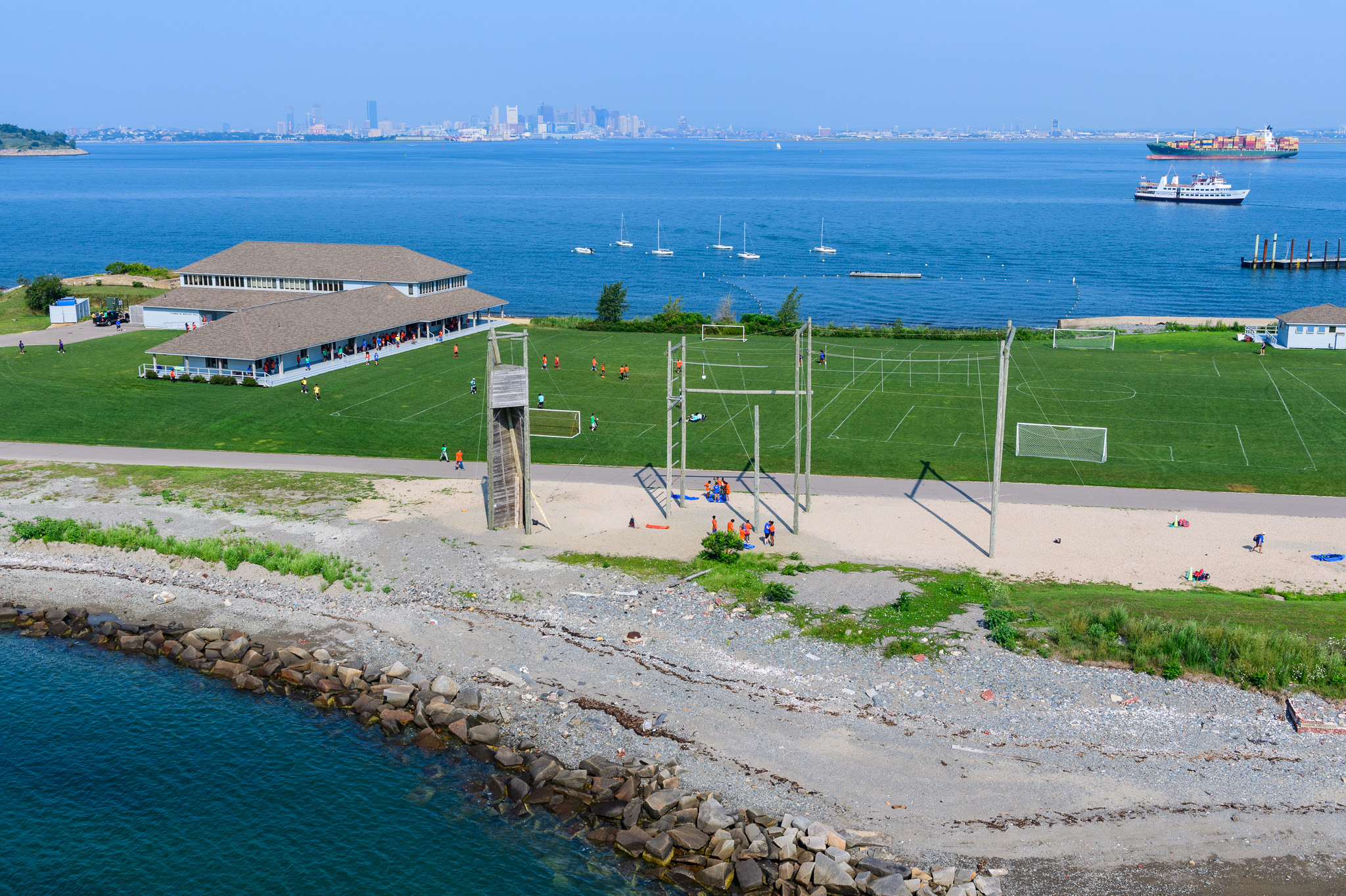 Camp Harbor View from the air, overlooking a green soccer field, a ropes course, and pavilion. The camp is surrounded by water and the Boston skyline is in the distance.