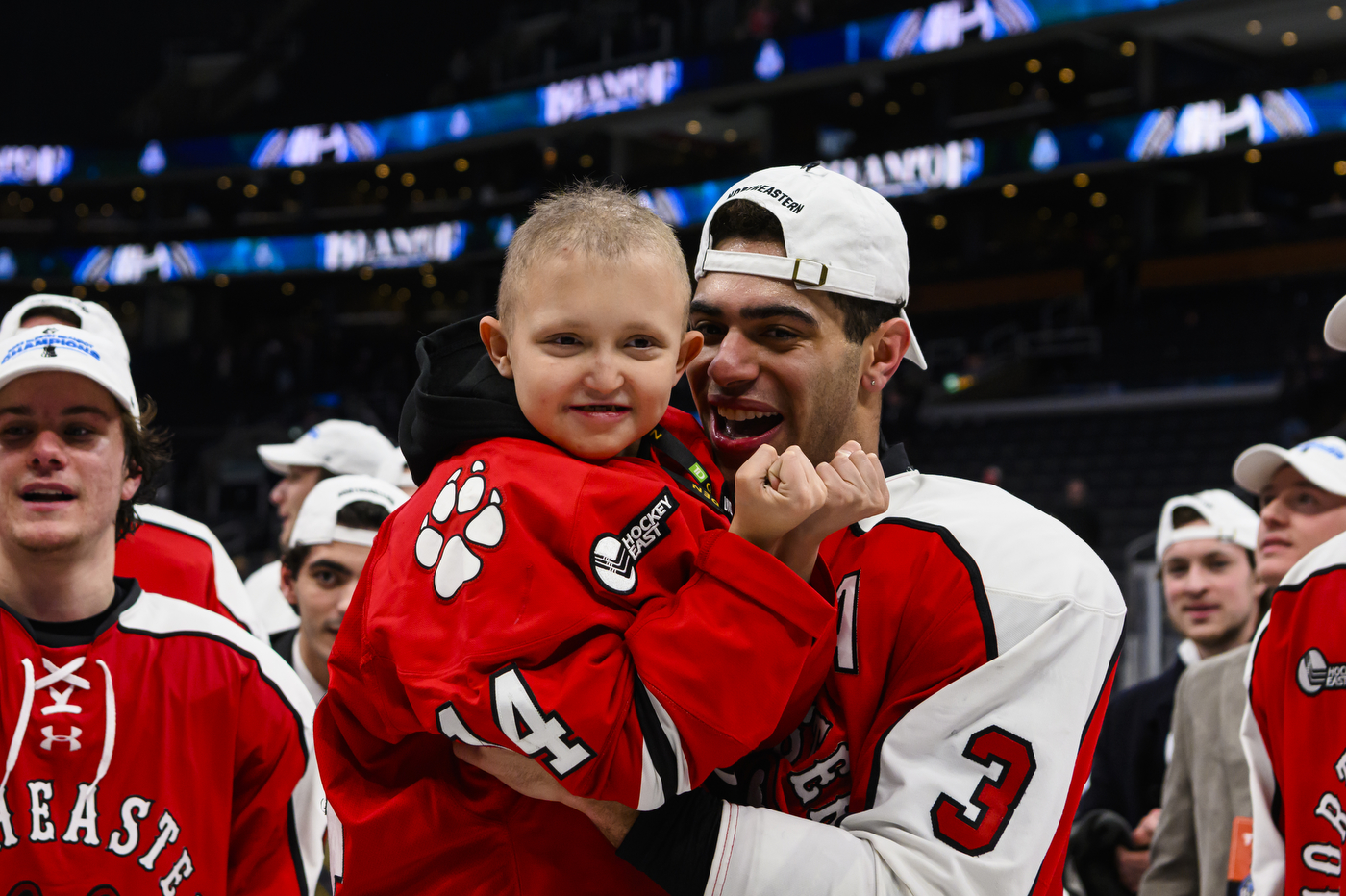 The Northeastern Men's Hockey team celebrating their win, holding up a small child wearing their jersey