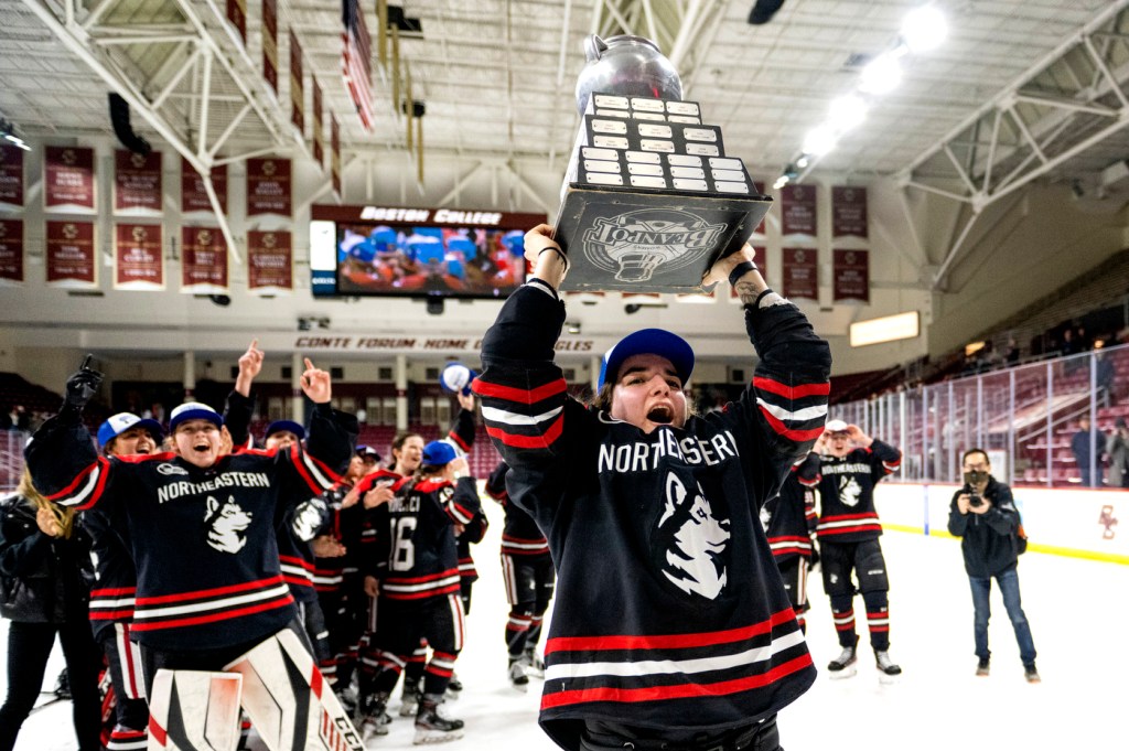 A Northeastern women's hockey player hoists the Beanpot tropy over her head and smiles as her teammates look on and cheer.