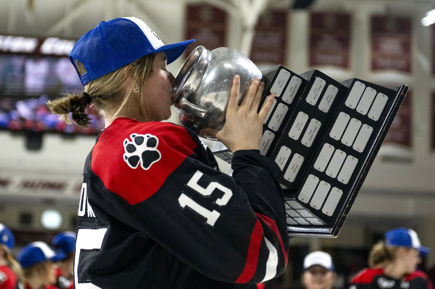 A hockey player dressed in a game jersey and a baseball hat kisses the Beanpot trophy