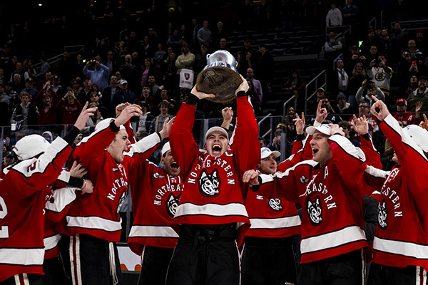 A Northeastern men's hockey player hoists the Beanpot trophy over his head and smiles as his teammates look on and cheer.