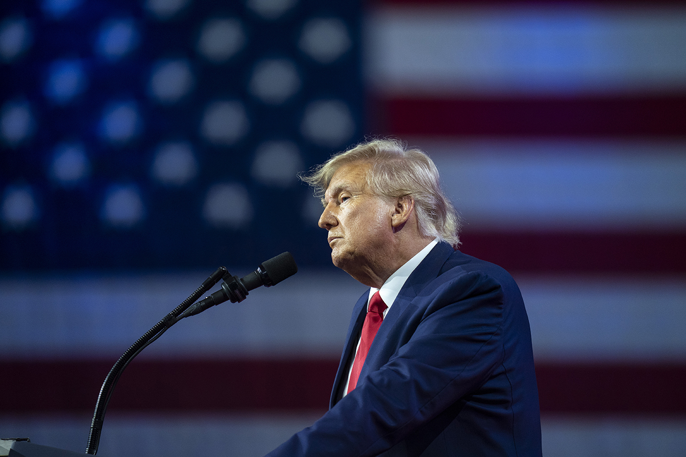 former President Donald Trump speaking into a microphone with a US flag hanging in the background