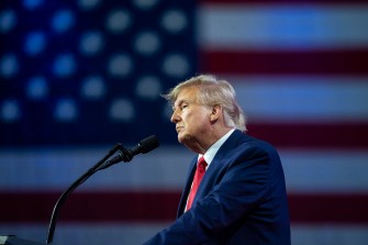 former President Donald Trump speaking into a microphone with a US flag hanging in the background