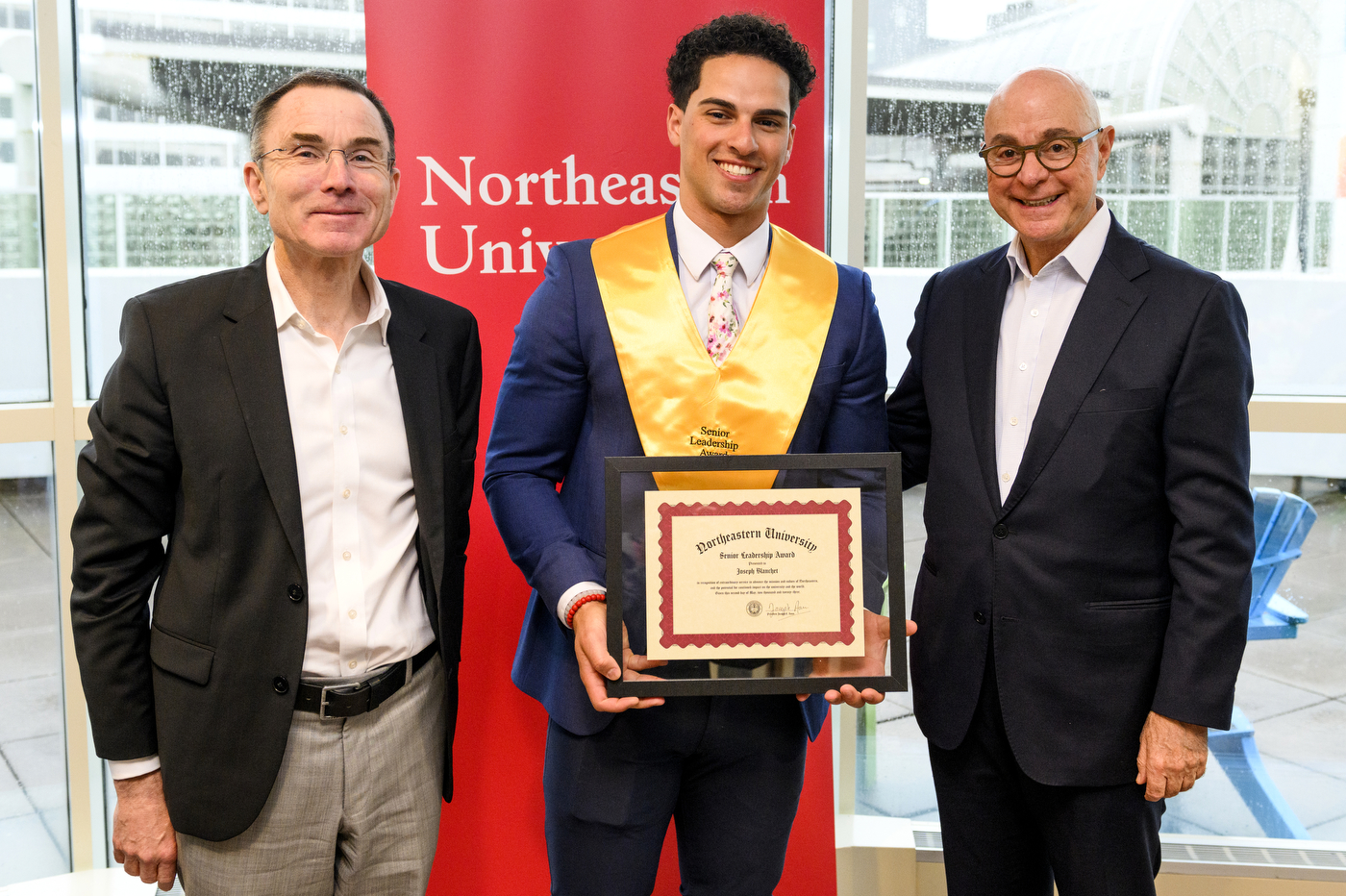 Northeastern student wearing gold sash and holding certificate next to President Aoun