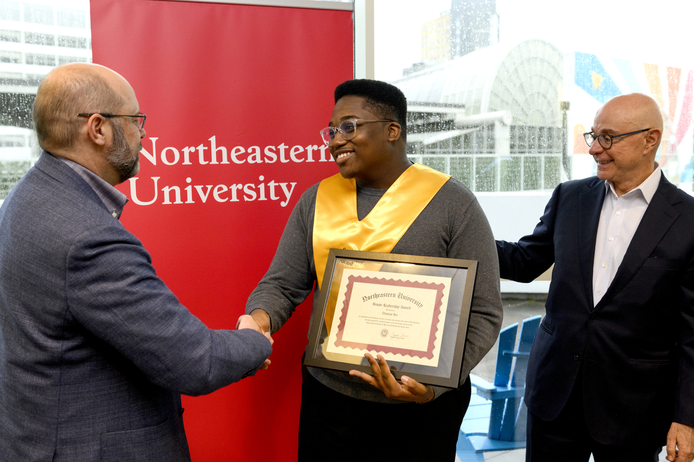 Northeastern student wearing gold sash and holding certificate next to President Aoun