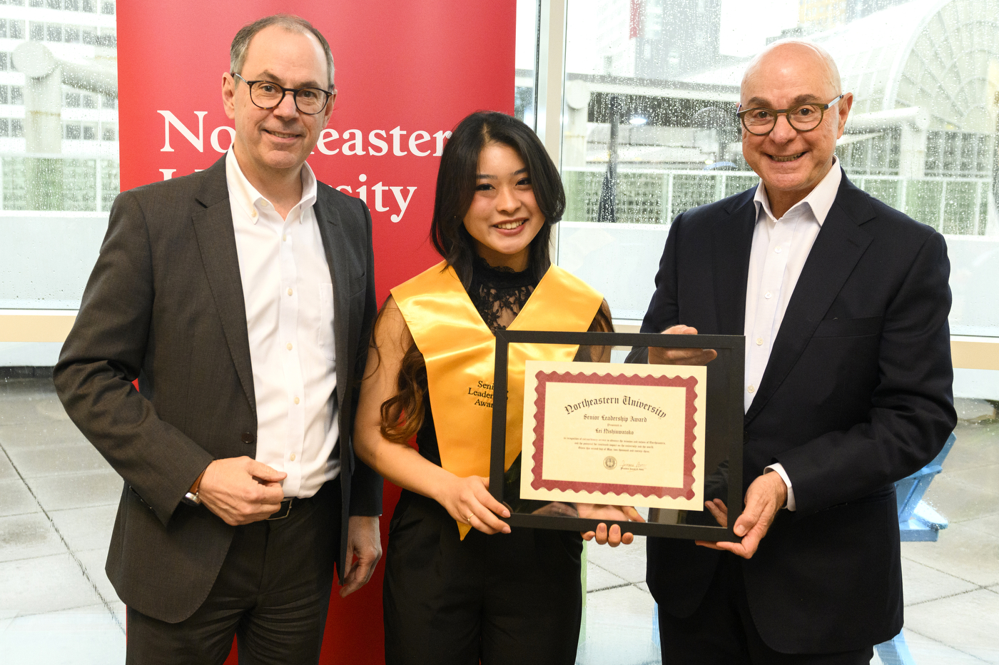 Northeastern student wearing gold sash and holding certificate next to President Aoun