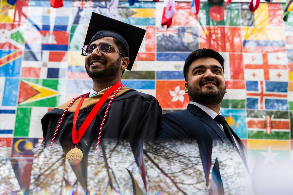 Two students, including one in cap and gown, pose against a backdrop of international flags