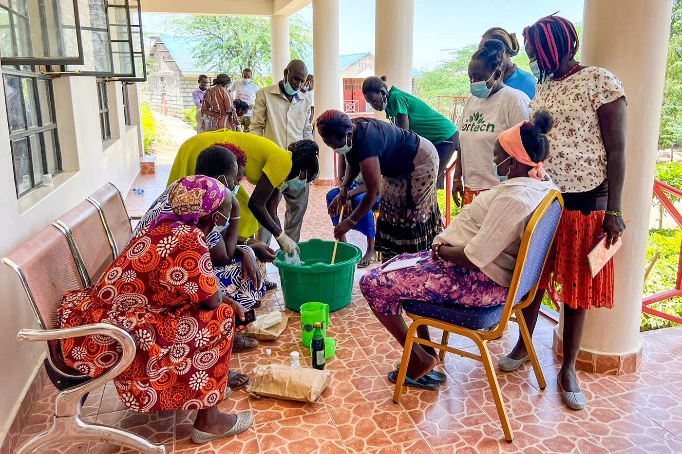 Multiple people sit and stand outside around a green bucket.