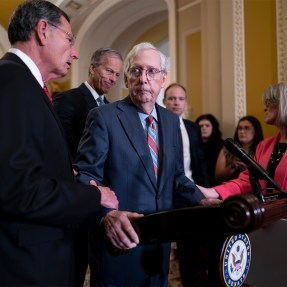 Mitch McConnell stands behind a podium at a news conference, while John Barrasso and Joni Ernst hold onto his wrists. Other officials stand behind Mitch McConnell.