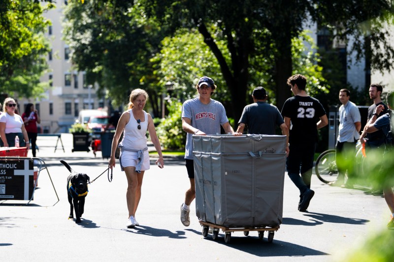 student pushing a wheeled bin of their belongings while a family member walks a dog next to them