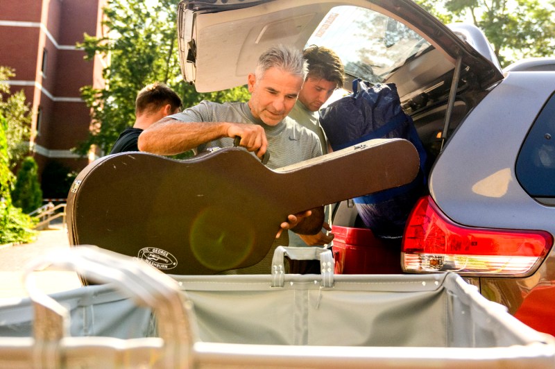 A parent helps a student load a guitar from a car trunk into a rolling bin