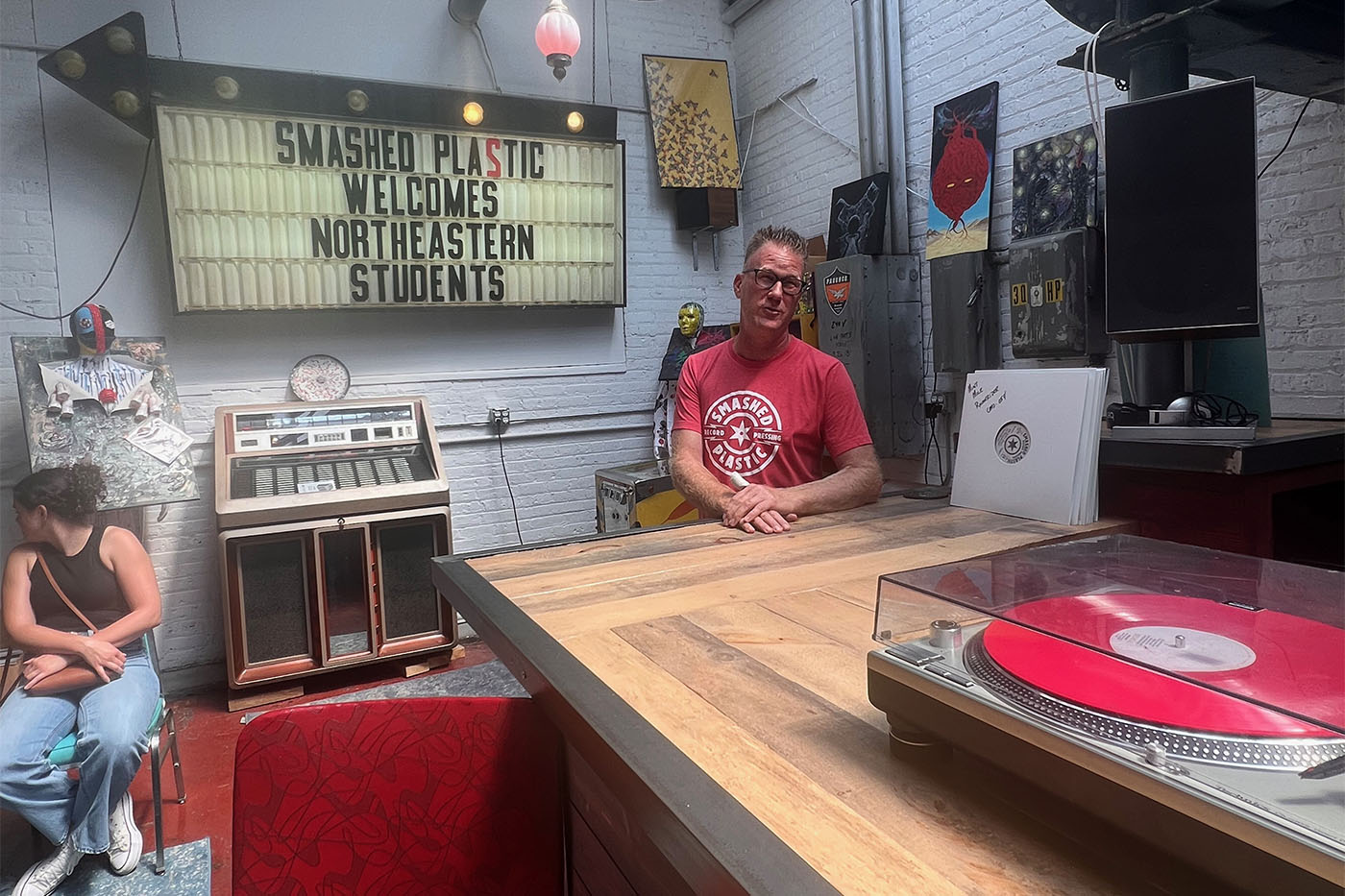 Person sitting at a large wooden table in front of a sign that reads "Smashed Plastic Welcomes Northeastern Students".