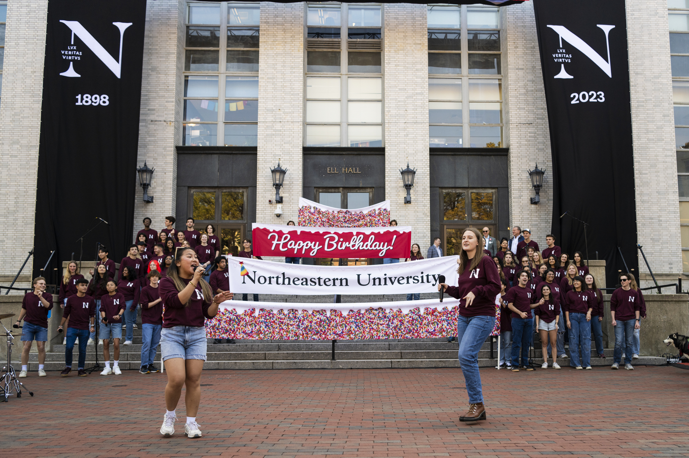 Two students speaking into microphones before Happy Birthday banners outside of Ell Hall.