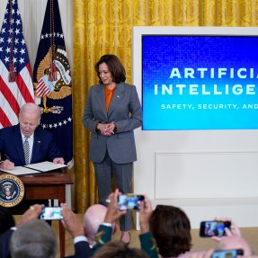 Kamala Harris standing over President Biden as he signs a new executive order on AI in the White House.