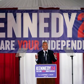 RFK Jr speaking during a campaign event with a banner behind him that reads 'KENNEDY24 DECLARE YOUR INDEPENDENCE' in all caps.