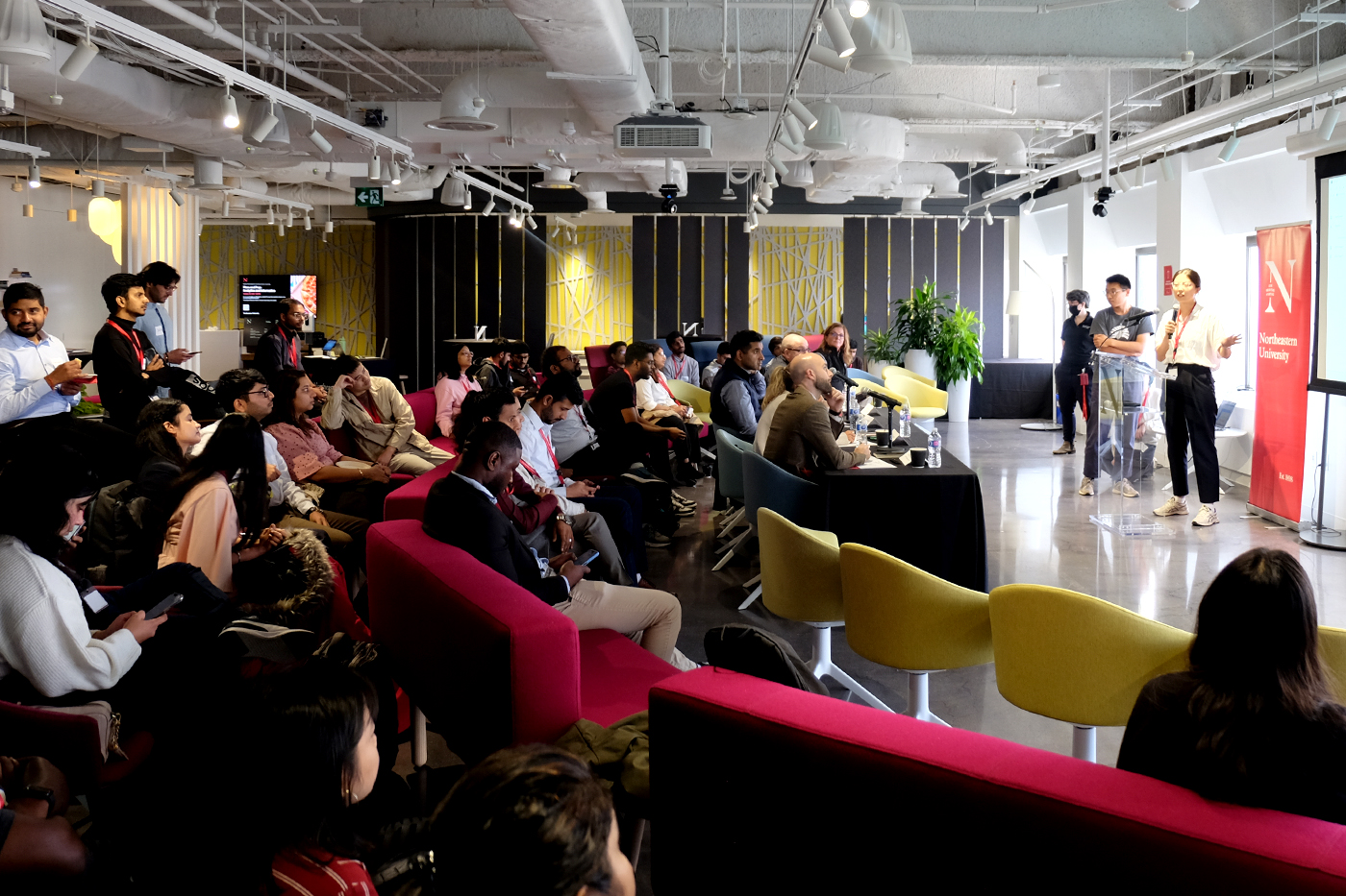 Students sitting in a room on red and yellow chairs listening to a speaker at Northeastern's Walmart Hackathon.