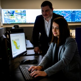 Alyssa Berg and JJ Cappa working on a laptop in an office with many screens.