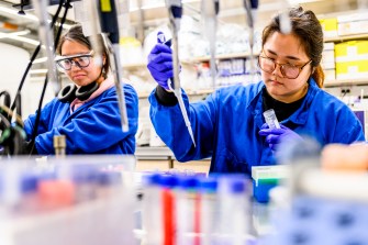 Two students dressed in protective gear, including gloves, use test tubes in a lab.