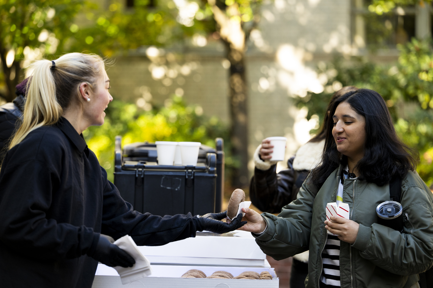 A person hands another person a donut outside on a sunny day.