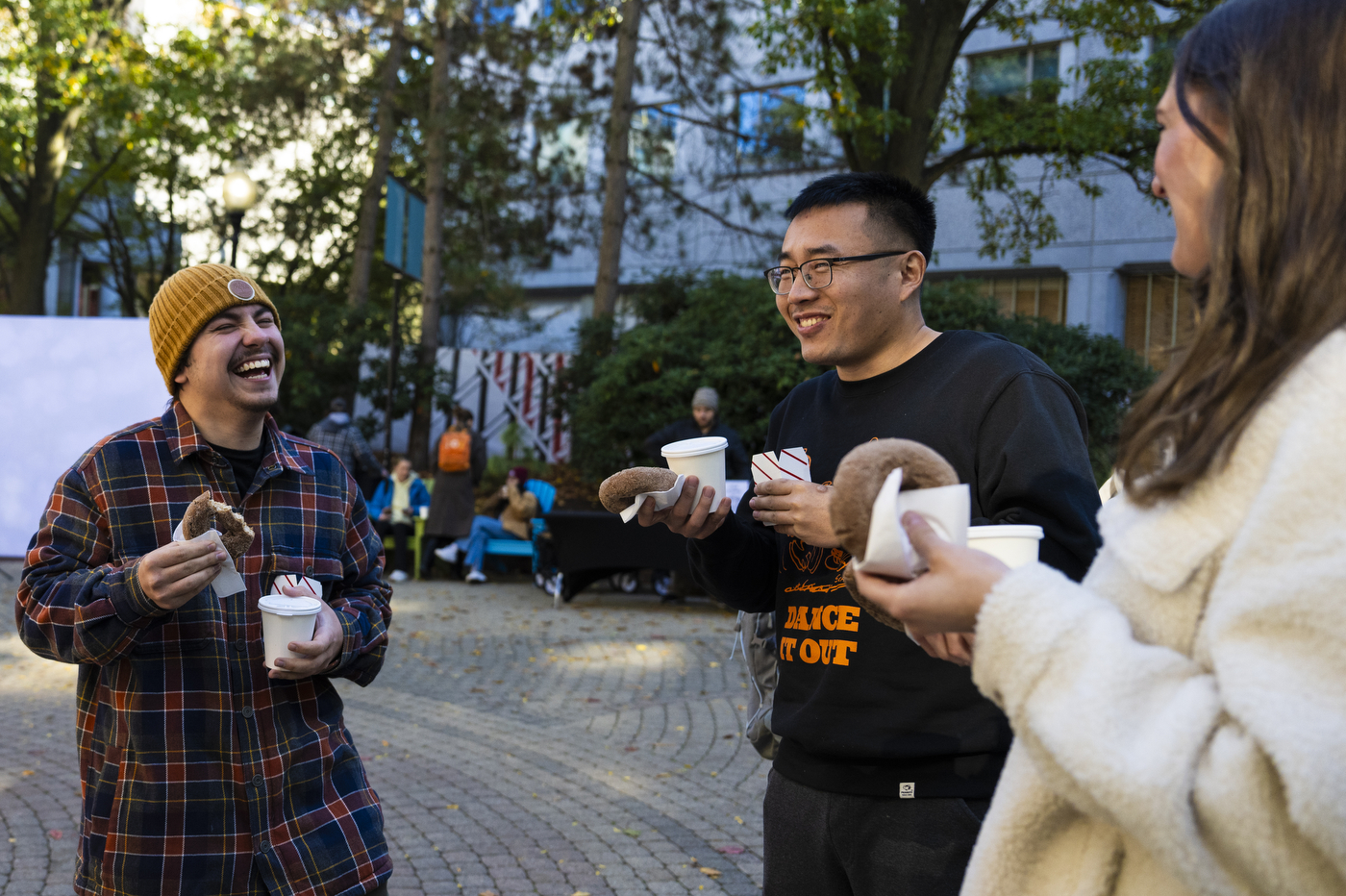 Three people laugh and eat donuts outside on a sunny day.